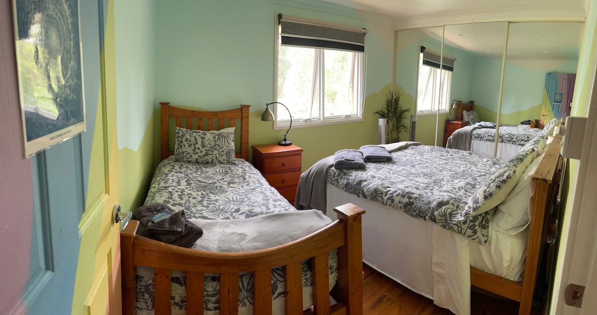 The bedroom features two beds, one spacious and one single, both adorned with floral-patterned bedding. A bedside table with a lamp beside each bed is visible. Natural light floods the room through large windows, complemented by mirrored wardrobes on one wall.