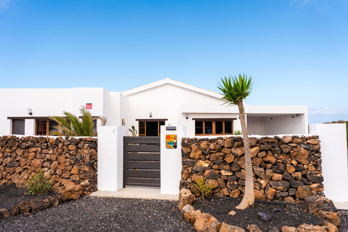 A modern villa entrance is framed by a stylish stone wall with a wooden gate. The exterior features white walls, a sloped roof, and tropical plants, creating a welcoming atmosphere. The clear blue sky adds to the serene setting.