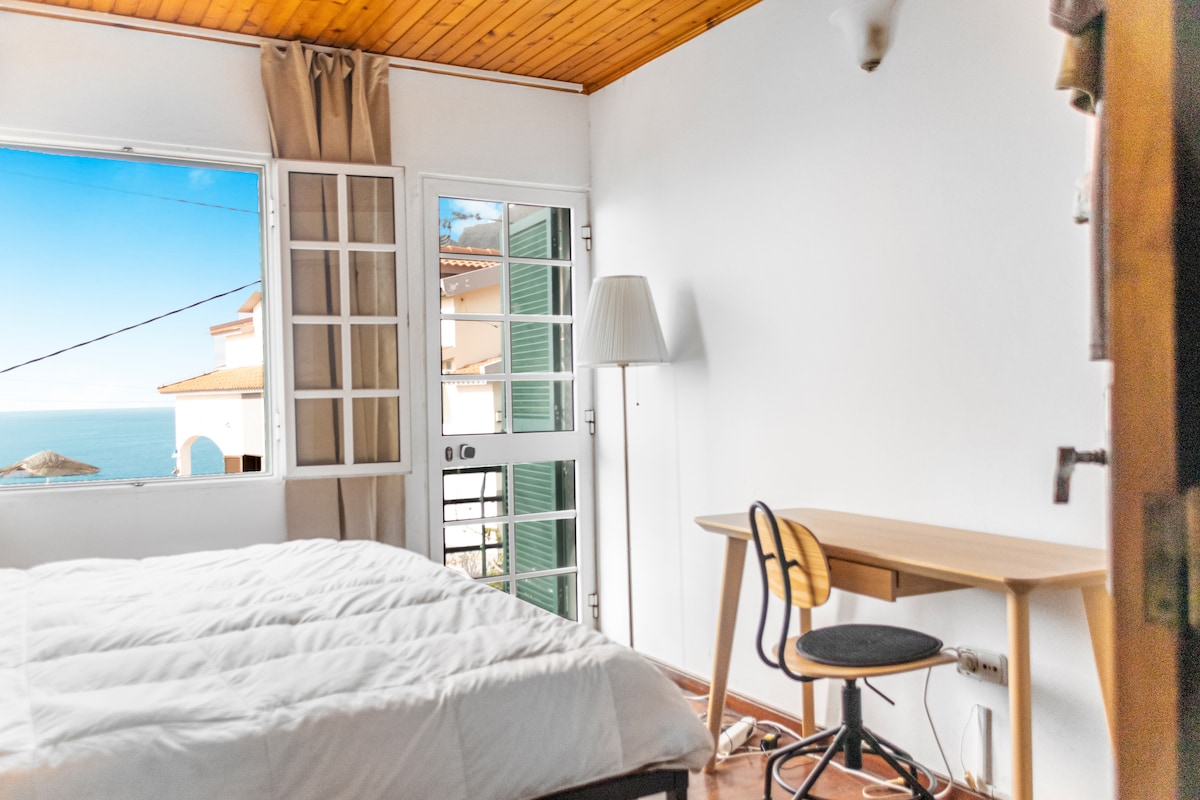 A bright room features a spacious bed, a desk with an ergonomic chair, and a large window offering views of the Atlantic Ocean. Natural light illuminates the space, and a standing lamp adds a practical touch. Doors lead to a private balcony.