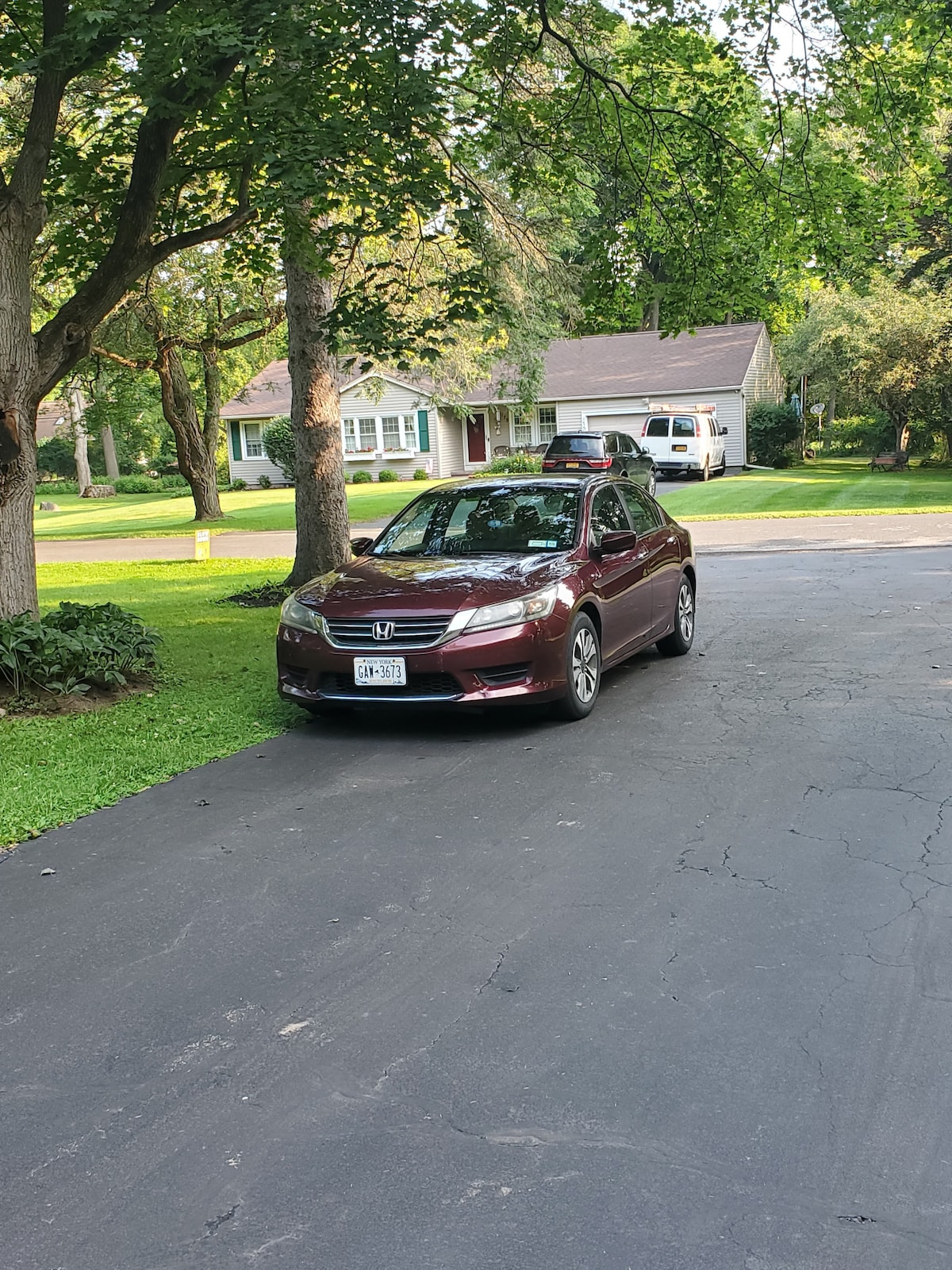 A burgundy sedan is parked on a black asphalt driveway surrounded by green grass. Trees provide shade in the area, and a single-story house is visible in the background along with another vehicle. The scene reflects a quiet, residential neighborhood.