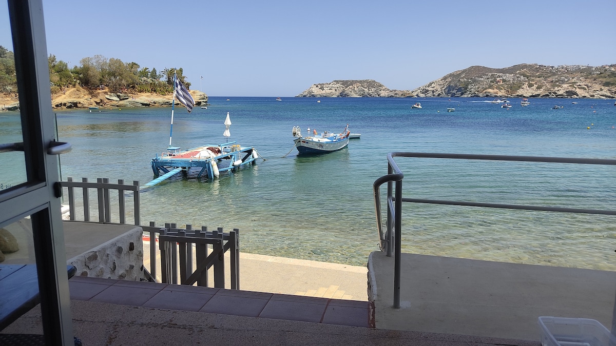 A serene view of calm blue waters is presented, with fishing boats anchored nearby. The shoreline is visible, leading to a distant rocky landscape, with greenery lining the coast. The image captures a beachside scene under clear skies, inviting relaxation.