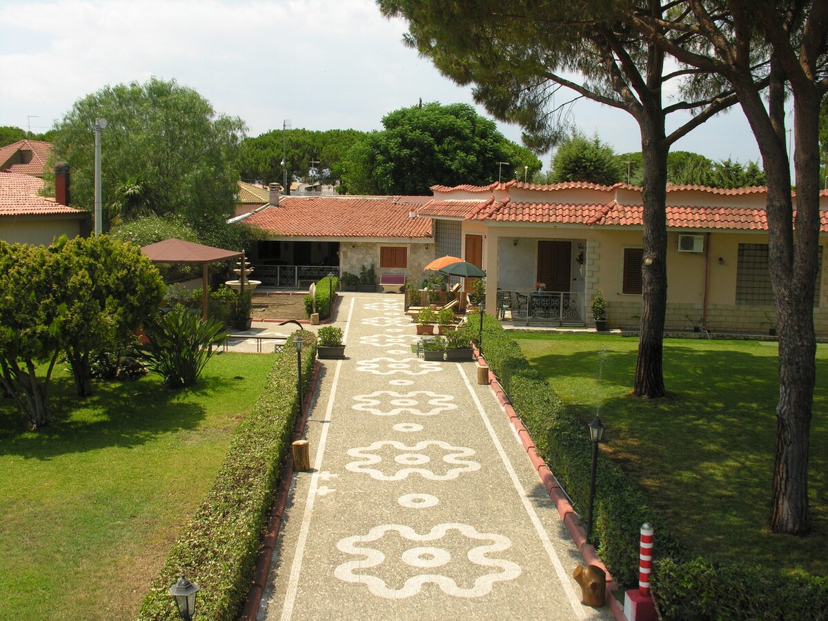 A charming pathway is bordered by manicured lawns and neatly arranged gardens, leading towards a villa with a terracotta roof. Outdoor seating areas and a shaded gazebo are evident, creating an inviting space for relaxation surrounded by greenery.