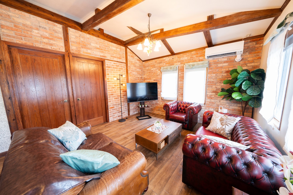 The living area features exposed brick walls and wooden beams, creating a cozy atmosphere. Two leather sofas offer ample seating, accompanied by a wooden coffee table. A television is positioned for easy viewing, while large windows allow natural light to fill the space.