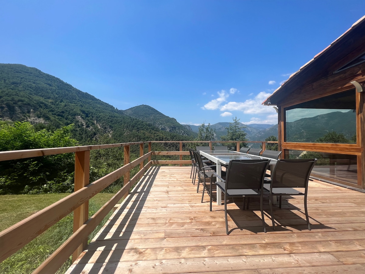 A spacious wooden terrace features a long dining table surrounded by black chairs, offering stunning views of the mountains and lush greenery. Natural light enhances the inviting atmosphere, and the surrounding landscape can be seen against a clear blue sky.