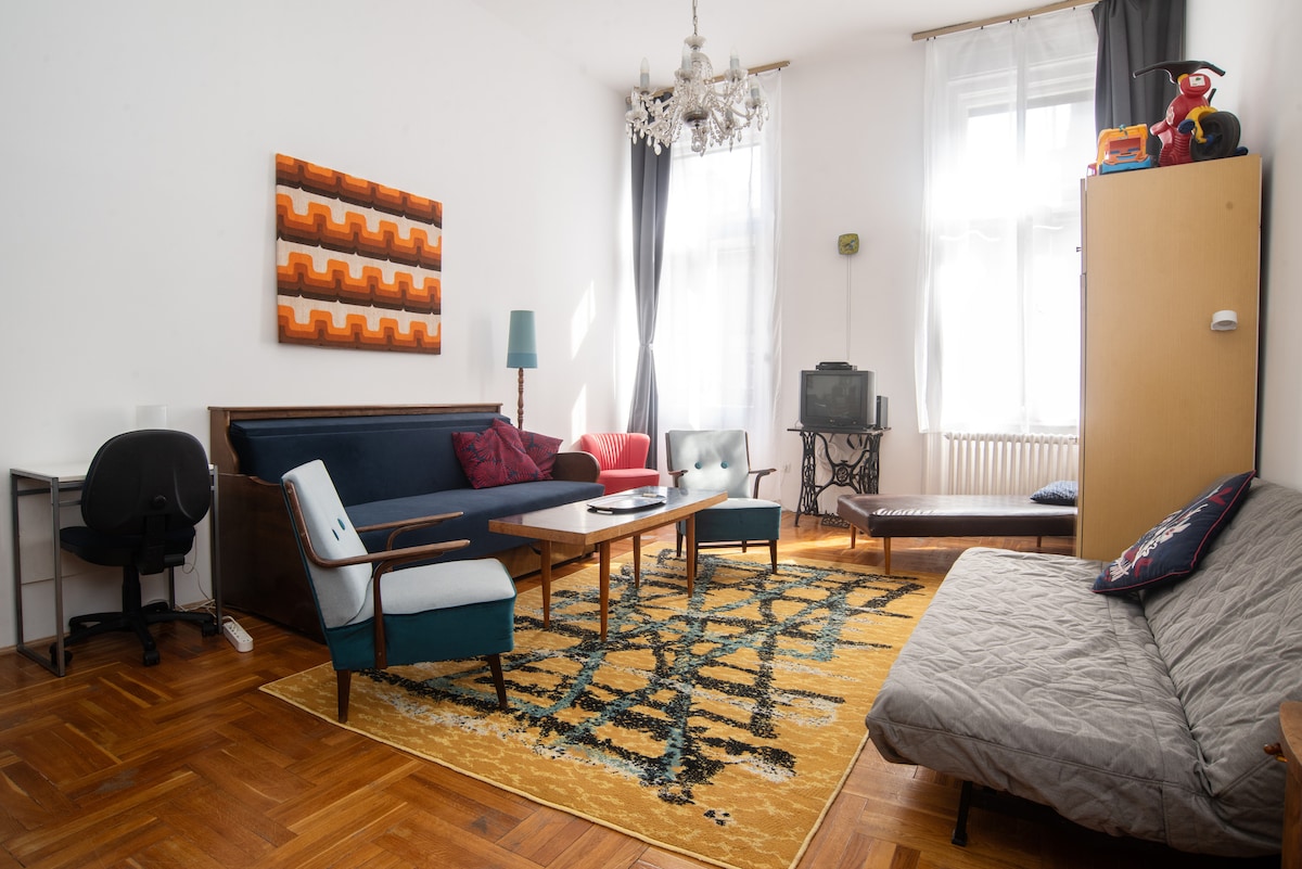 The living room showcases a combination of retro-inspired furnishings, including a large rug with geometric patterns. Two pull-out sofas are positioned around a central table, with ample natural light from big windows. A vintage chandelier hangs above, creating an inviting space complemented by decor accents.