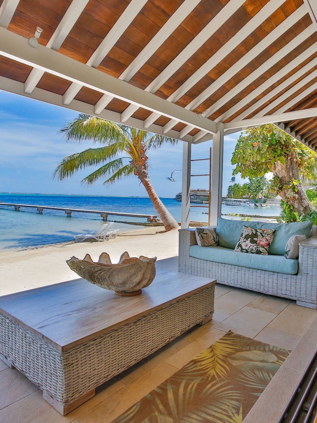 An outdoor seating area features a woven coffee table and comfortable seating adorned with tropical-patterned cushions. The space opens up to a sandy beach, framed by palm trees and the blue lagoon in the background. A private dock is visible in the distance.