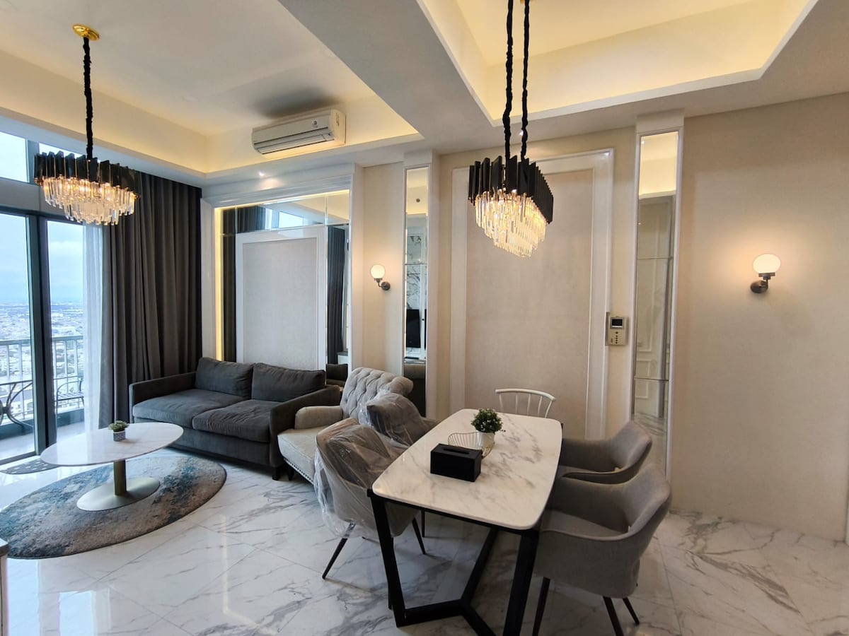 A modern living space is visible, featuring a comfortable gray sofa and a stylish marble dining table surrounded by elegant chairs. Soft lighting fixtures hang from the ceiling, enhancing the contemporary feel. Large windows provide a view of the surrounding cityscape.
