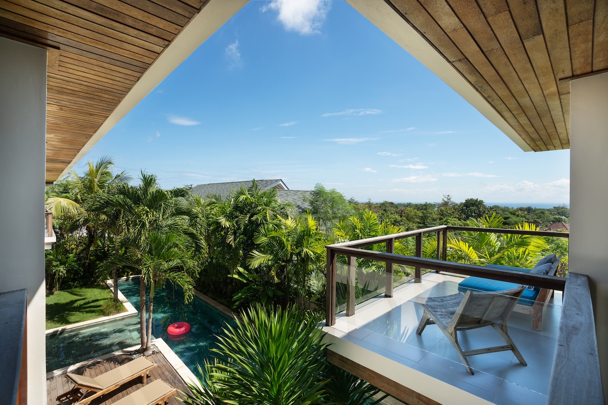 A serene balcony view captures a tropical landscape, featuring lush greenery and a swimming pool. A wooden chair and a beach towel are positioned on the balcony, inviting relaxation. The blue sky overhead complements the vibrant colors of the surrounding plants.