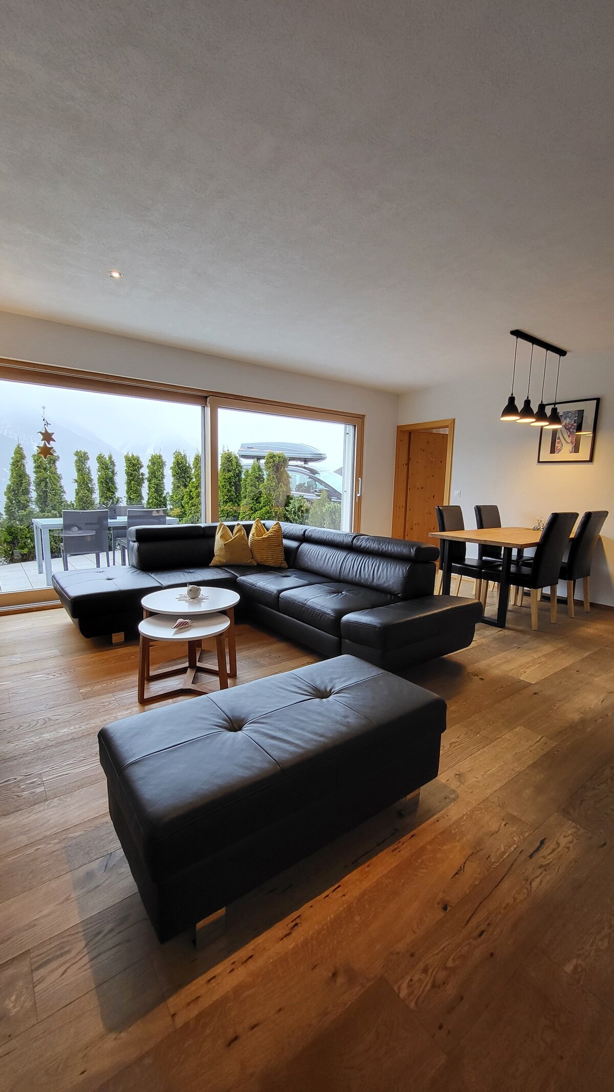 A spacious living area features a sleek black leather sectional sofa and a small round table. Large windows provide natural light and a view of the outdoor terrace and surrounding greenery. A dining table with seating for four is visible, complementing the modern decor.