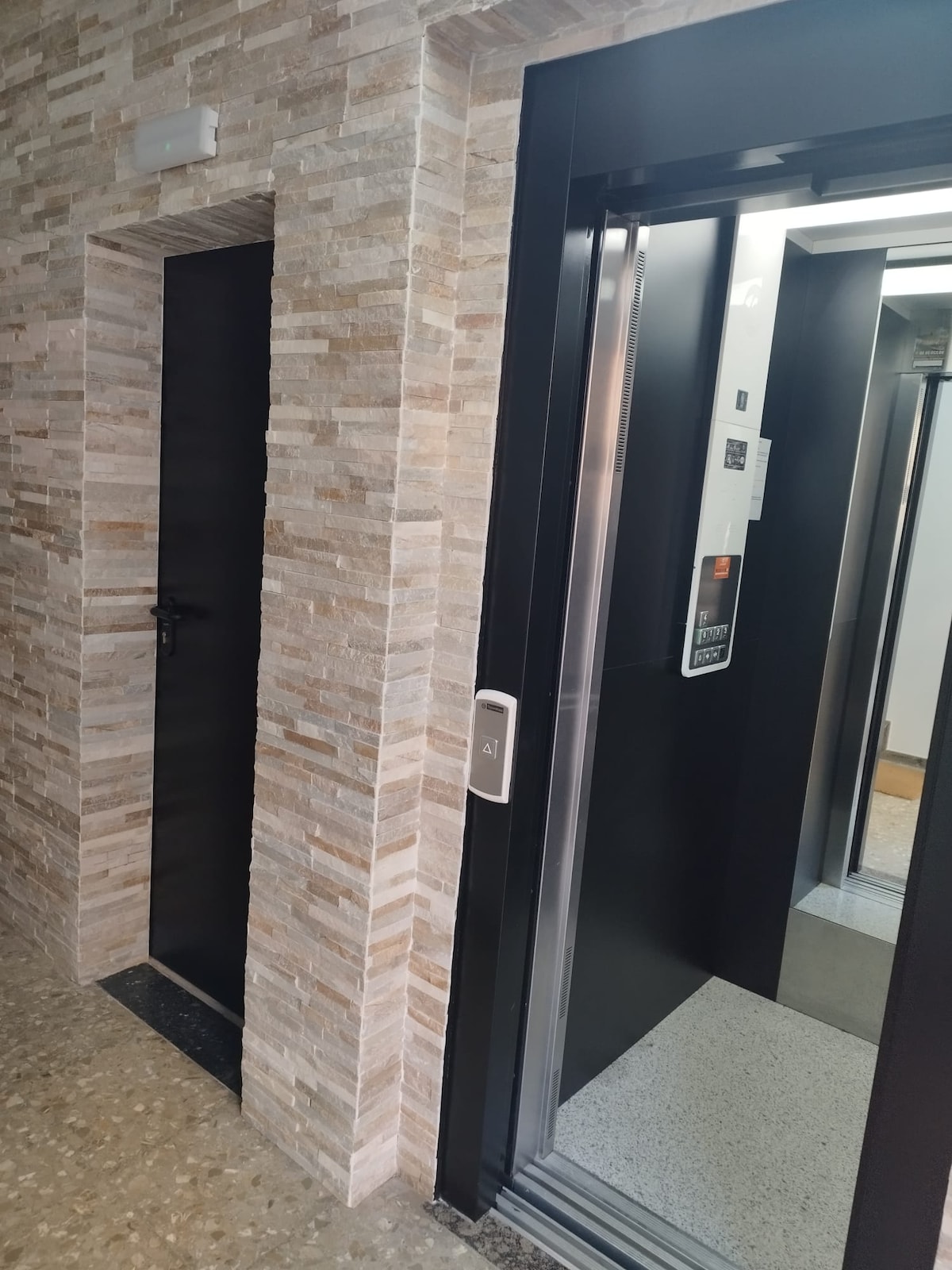 An elevator is located next to a door framed by light-colored stone walls. The dark door contrasts with the surrounding wall, which displays a textured finish. The elevator features metallic panels and a digital display, providing access to upper floors.