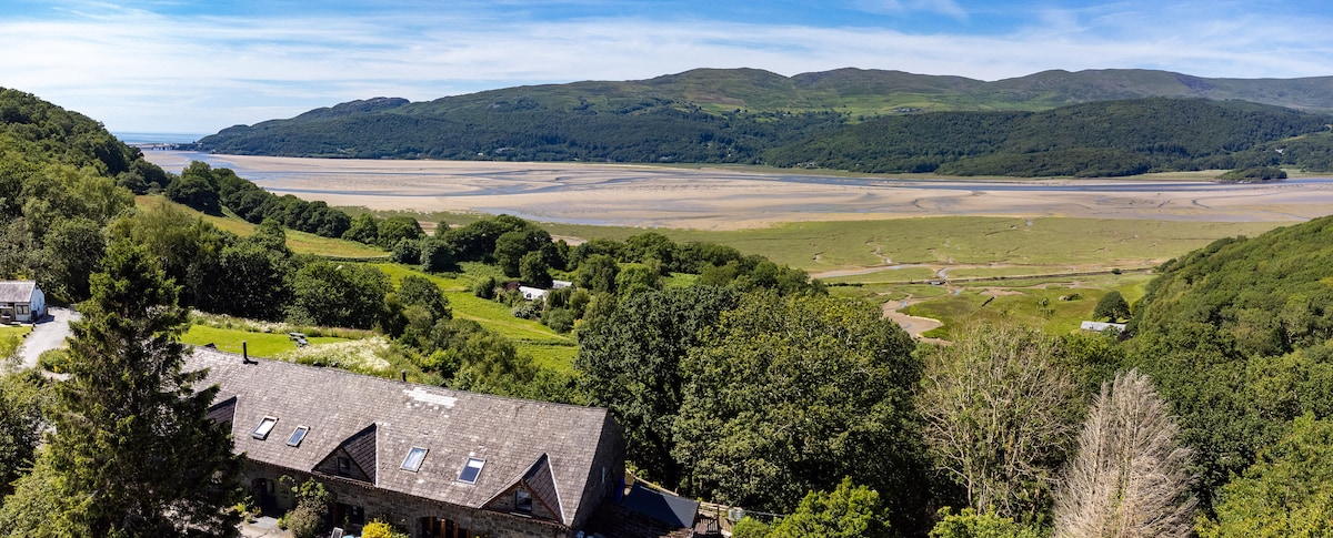 A panoramic view captures the expansive Mawddach Estuary, flanked by lush green hills and woodlands. The foreground features a cottage with a distinctive slate roof, blending harmoniously into the natural landscape, which stretches across the horizon under a bright blue sky.
