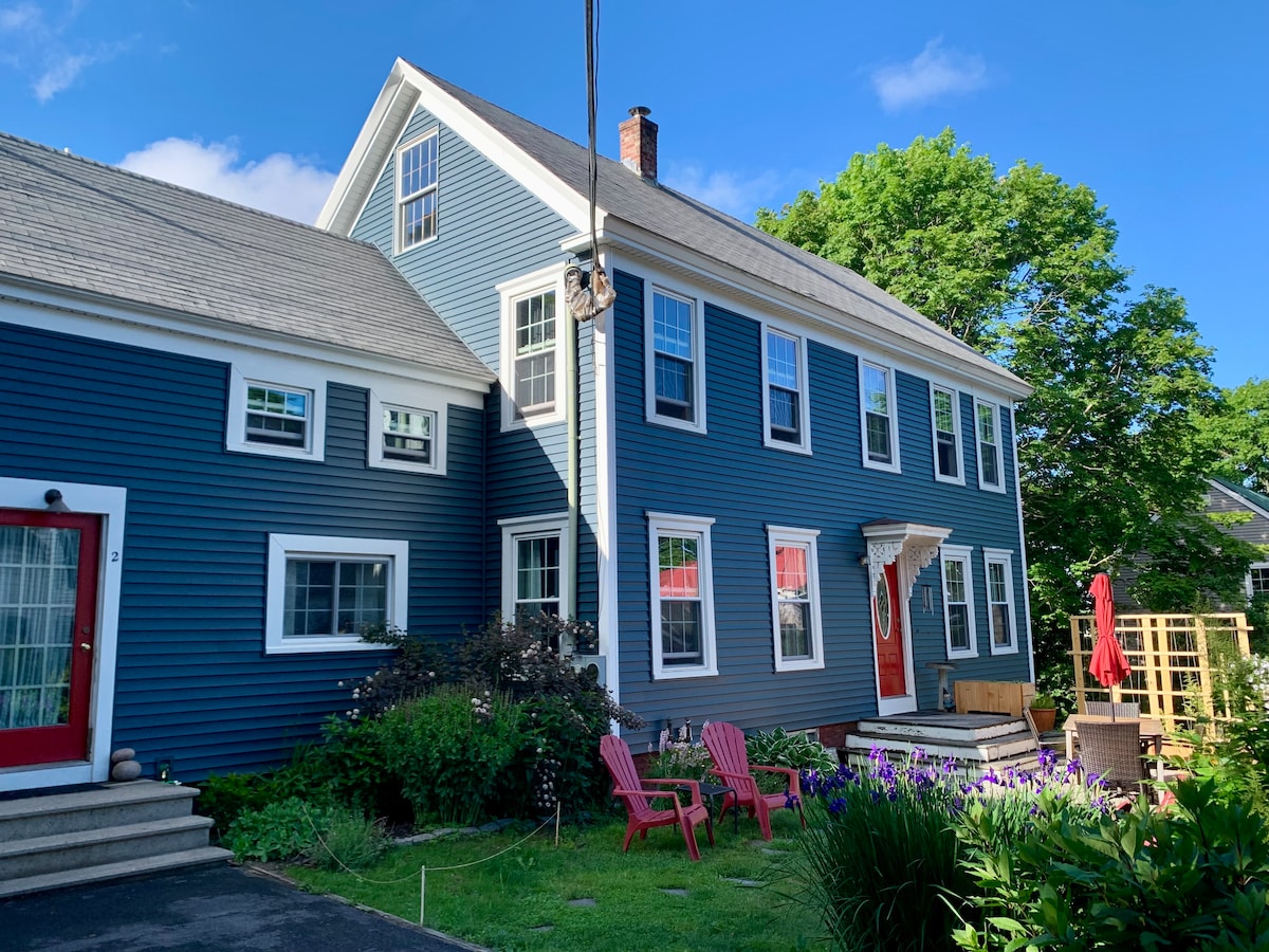 A charming family-sized home is showcased with blue siding and white trim. Adirondack chairs sit invitingly on the lawn, surrounded by vibrant greenery. The well-maintained garden features colorful flowers, and a patio is visible with a red umbrella, enhancing the outdoor gathering space.