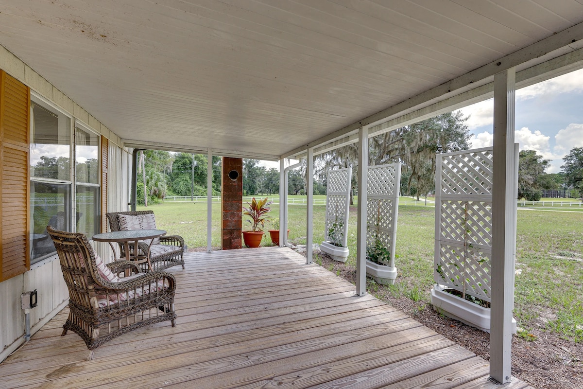 A spacious covered porch is featured with a wooden floor and two comfortable chairs. The surrounding area showcases a view of the expansive green yard, providing a relaxing outdoor space near nature.