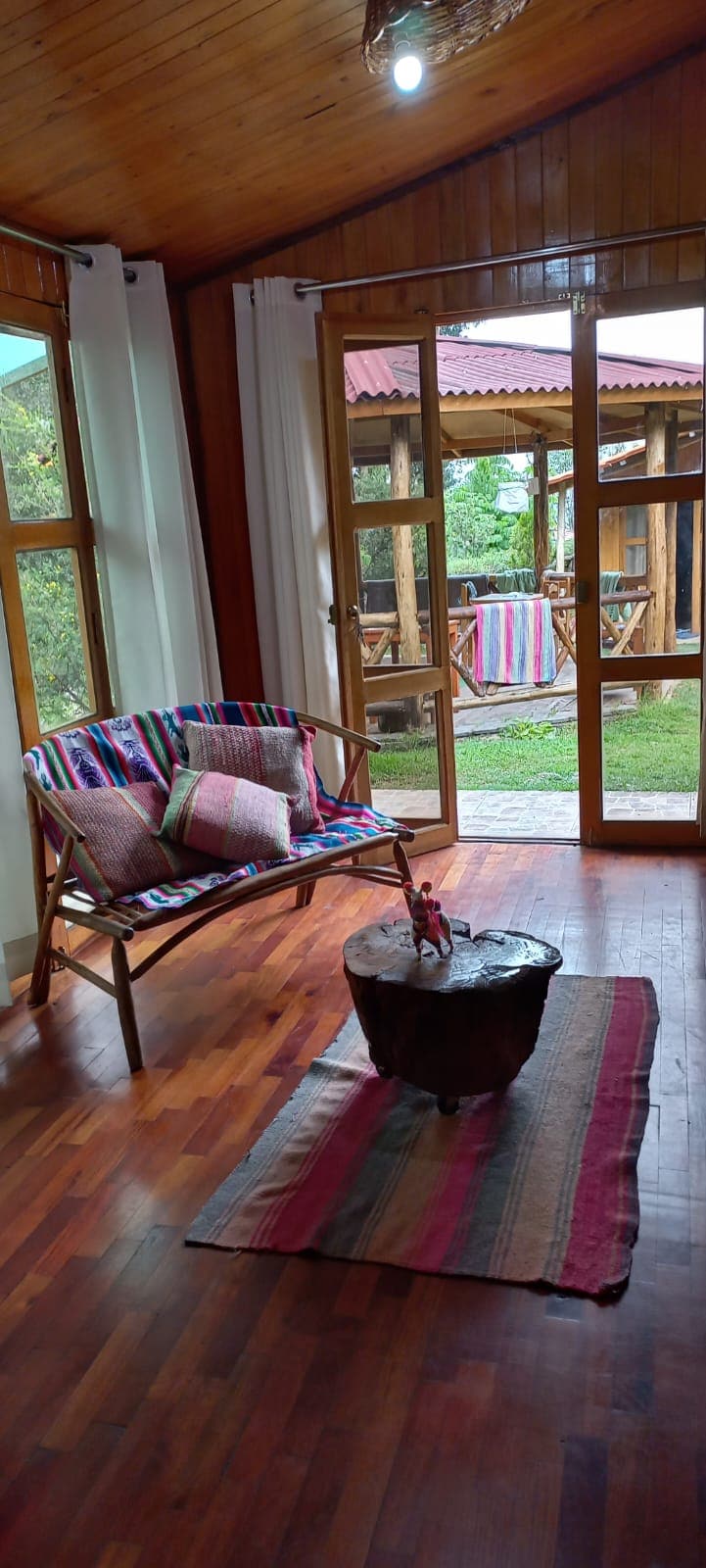 A cozy living area features a wooden floor and large windows allowing natural light to fill the space. A colorful, striped chair with decorative cushions is positioned next to a round wooden coffee table, enhancing the welcoming atmosphere. Views of the garden area can be seen through the glass doors.