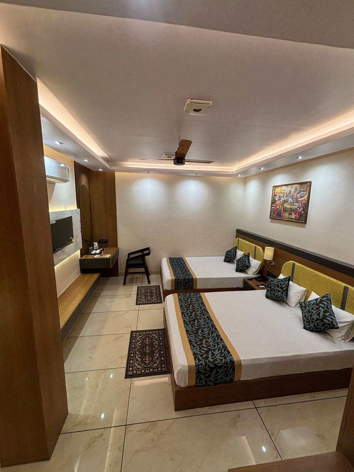 A well-appointed room features two spacious beds with decorative cushions and a patterned bed runner. A wall-mounted television is visible above a sleek wooden unit, while soft lighting enhances the clean, modern aesthetic. A chair and a small table complete the comfortable setting.