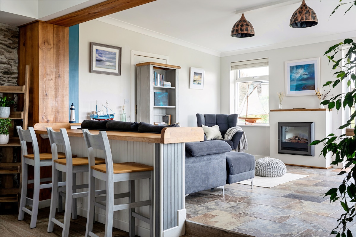 A spacious open-plan living area features a comfortable grey sofa, a cozy rug, and a modern electric fireplace. A wooden dining bar with seating for four is positioned beside the light-filled kitchen, complemented by calming coastal artwork on the walls and natural light from the windows.