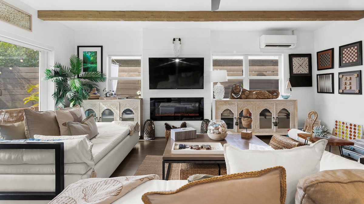 A modern living room features two large plush couches and a stylish coffee table at its center. A mounted flat-screen TV is above a contemporary fireplace, surrounded by an array of decorative elements, including artwork and greenery, enhancing the serene atmosphere.