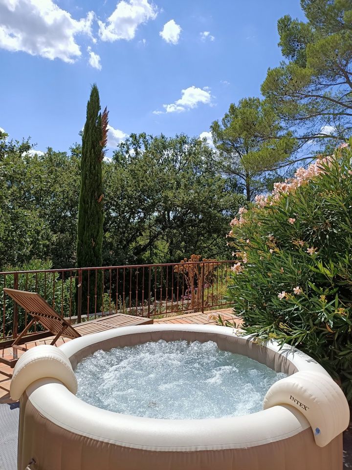 A private hot tub sits on a sunlit terrace, surrounded by lush greenery and flowering shrubs. Clear blue skies are visible overhead, with a single chair positioned nearby, providing a peaceful outdoor setting for relaxation.
