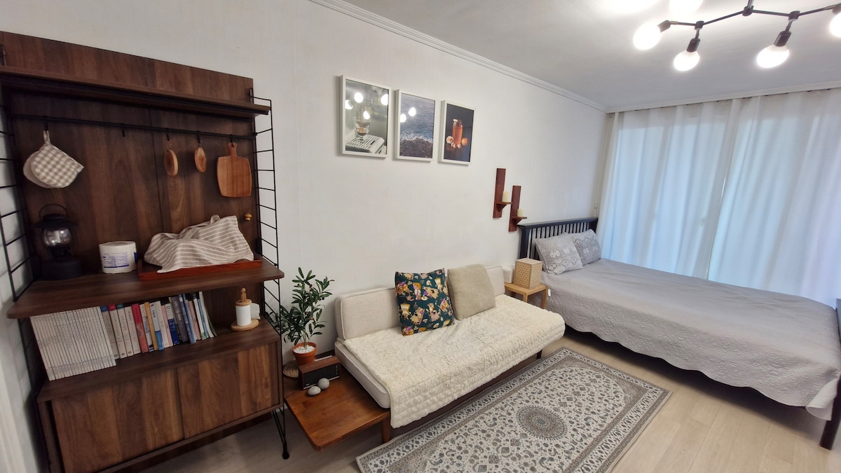 An open living space is presented, featuring a cozy sofa adorned with decorative pillows. A wooden shelf holds various items, including kitchen utensils and books. A spacious bed with a light grey cover sits against a wall, while soft natural light filters through sheer curtains.