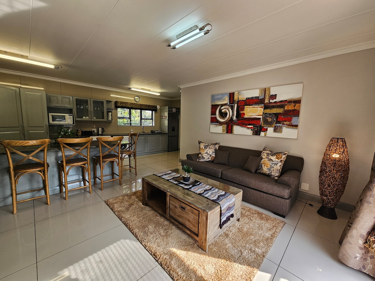 An open-plan living space features a comfortable seating area with a plush sofa, accompanied by a wooden coffee table. The kitchen is visible in the background, equipped with modern appliances and ample cabinetry. Decorative wall art adds a touch of colour to the neutral decor.
