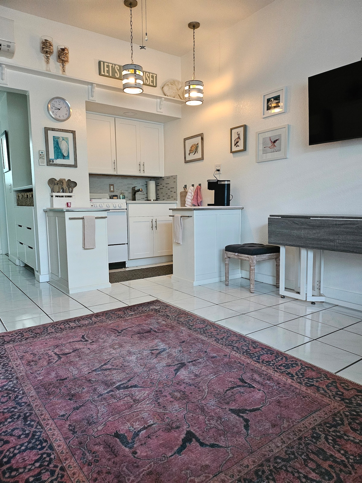 A compact kitchen area is visible, featuring white cabinetry and modern appliances. A small rug adds warmth, while decorative artwork enhances the space. A dining table with a gray bench is positioned nearby, and a flat screen TV is mounted on the wall.