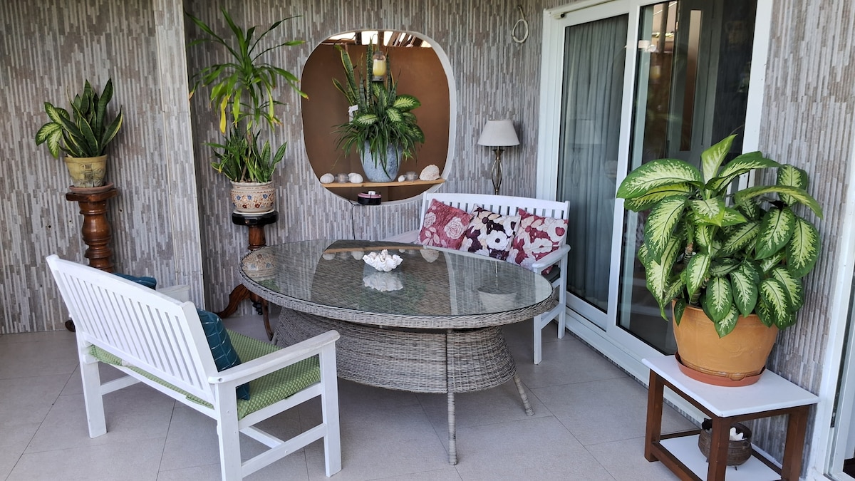 A cozy outdoor seating area is presented, featuring a circular glass table surrounded by comfortable seating. Numerous indoor plants add a touch of greenery, complemented by a decorative wall niche and a nearby lamp. Sliding glass doors provide access to the interior.