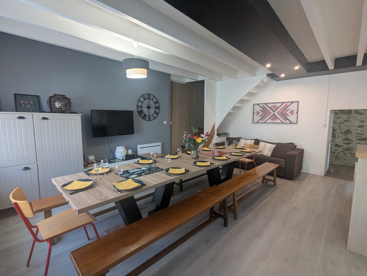 A spacious dining area is displayed, featuring a large wooden table with ample seating. Decorated in neutral tones, the room showcases a flat-screen TV mounted on the wall. A cozy seating area is seen in the background with a modern couch and decorative wall art.