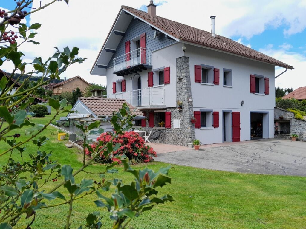 A two-story house with a light grey exterior and red shutters is set amidst a lush green lawn. The structure features a stone accent wall and multiple balconies. Vibrant flowering bushes enhance the outdoor space, offering a serene view of the surroundings.