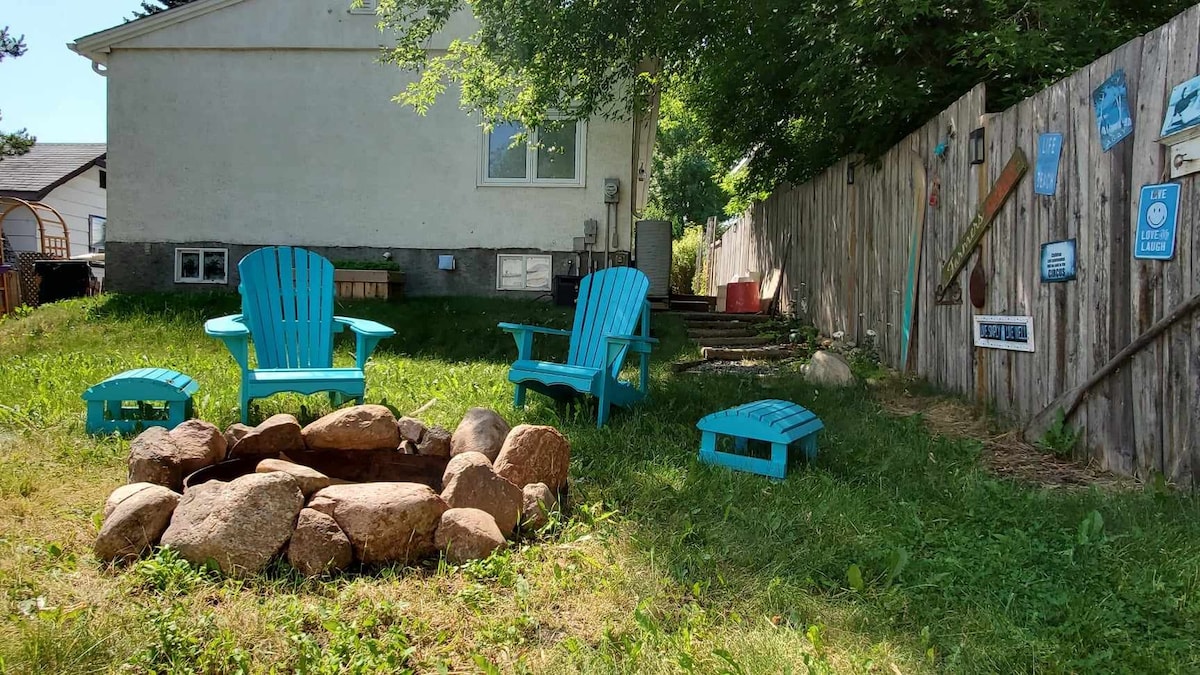 A backyard space features several turquoise adirondack chairs arranged around a natural stone fire pit. The well-kept grass provides a cozy setting, enhanced by a wooden fence partially adorned with decorative signs. Shade from overhanging trees adds a comfortable touch to the area.