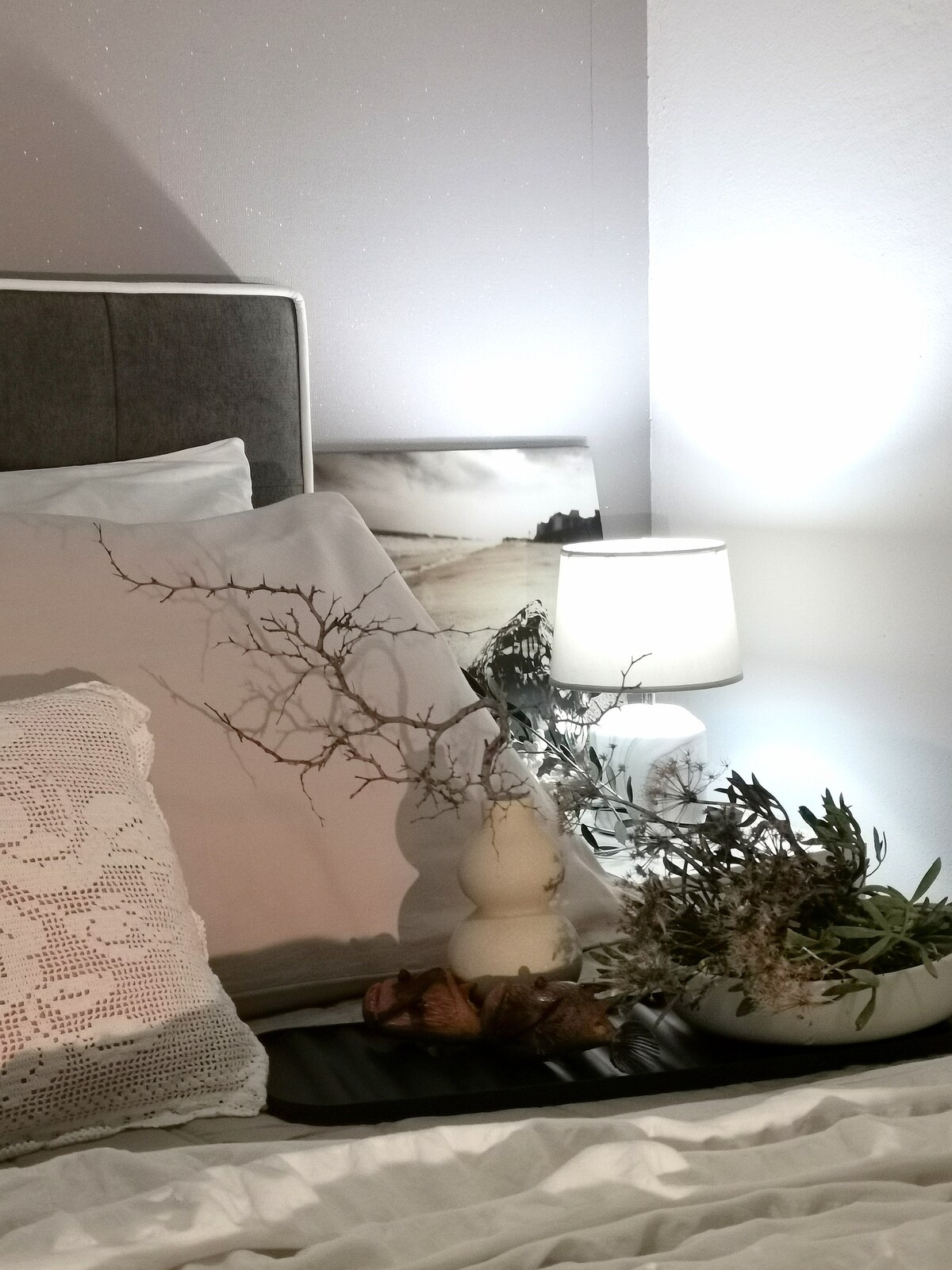 The bedroom features a neatly arranged bed with soft bedding and decorative pillows. A bedside table holds a lamp and a vase with a variety of natural elements, including plants and shells, creating a calming atmosphere.