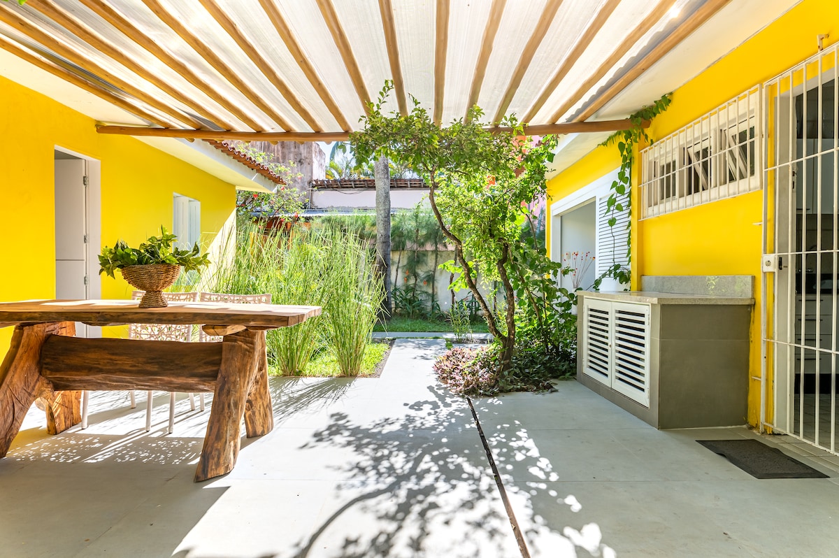 A spacious outdoor area is framed by vibrant yellow walls and shaded by a wooden pergola. A rustic wooden table is set in the center, surrounded by lush greenery and colorful plants, offering a serene space for relaxation and gatherings.