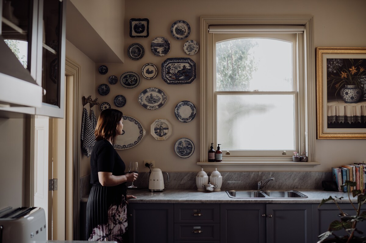 A well-appointed kitchen features a marble countertop and navy cabinets. Decorative blue and white plates adorn the wall above. A window allows natural light to fill the space, illuminating a woman who is standing beside the sink, holding a beverage and admiring the setup.