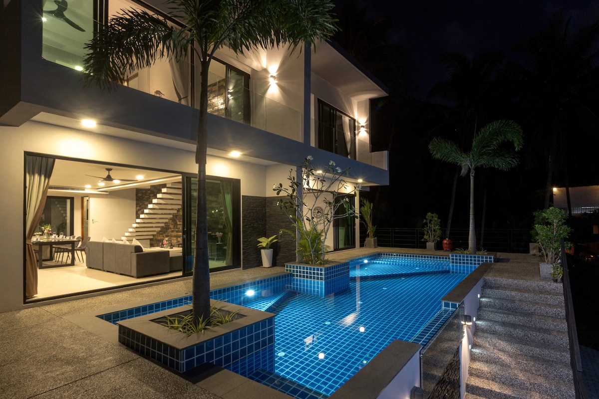 The modern villa is showcased at night, with a well-lit swimming pool reflecting the warm lights of the structure. The entrance features a landscaped area with tropical plants, while large windows enhance the view of the inviting living space and elegant stairs.