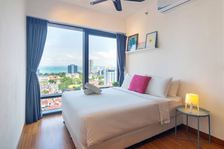 This modern bedroom features a luxurious King Koil bed, ample natural light from large windows showcasing the city and sea.