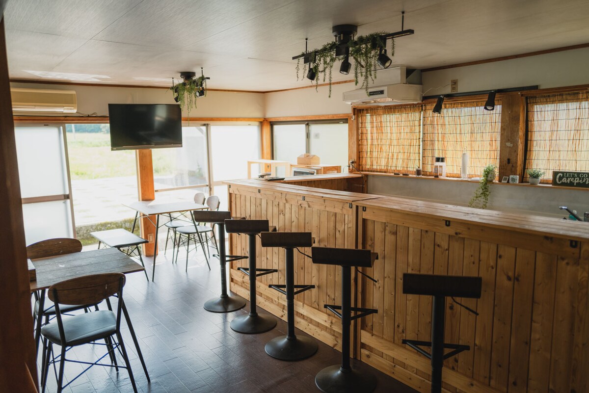 A spacious common area features a wooden bar with high stools and a counter, complemented by tables and chairs. Large windows provide natural light and a view of the surrounding landscape. Decorative plants hang from the ceiling, adding a fresh touch to the space.