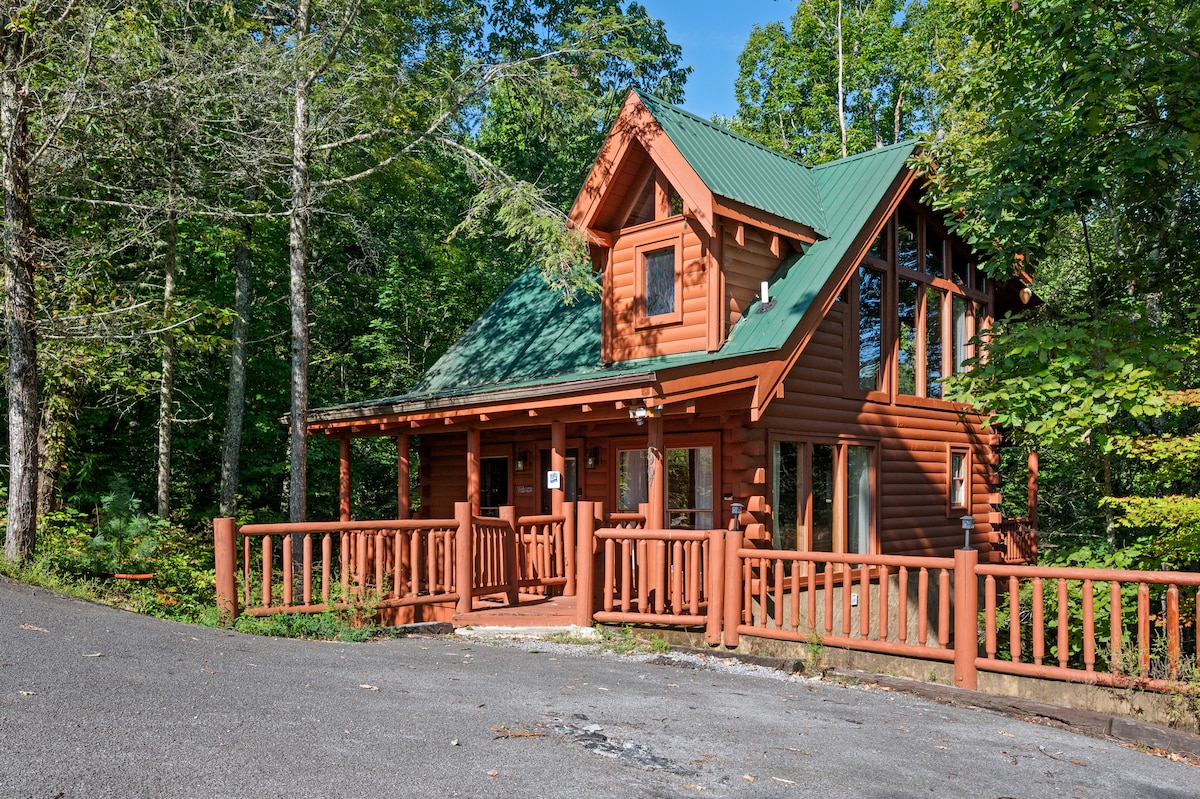 The classic log-style cabin features a striking orange-brown exterior with expansive windows and a green metal roof. A robust wooden deck wraps around the front, providing a welcoming entry surrounded by lush trees and natural greenery.