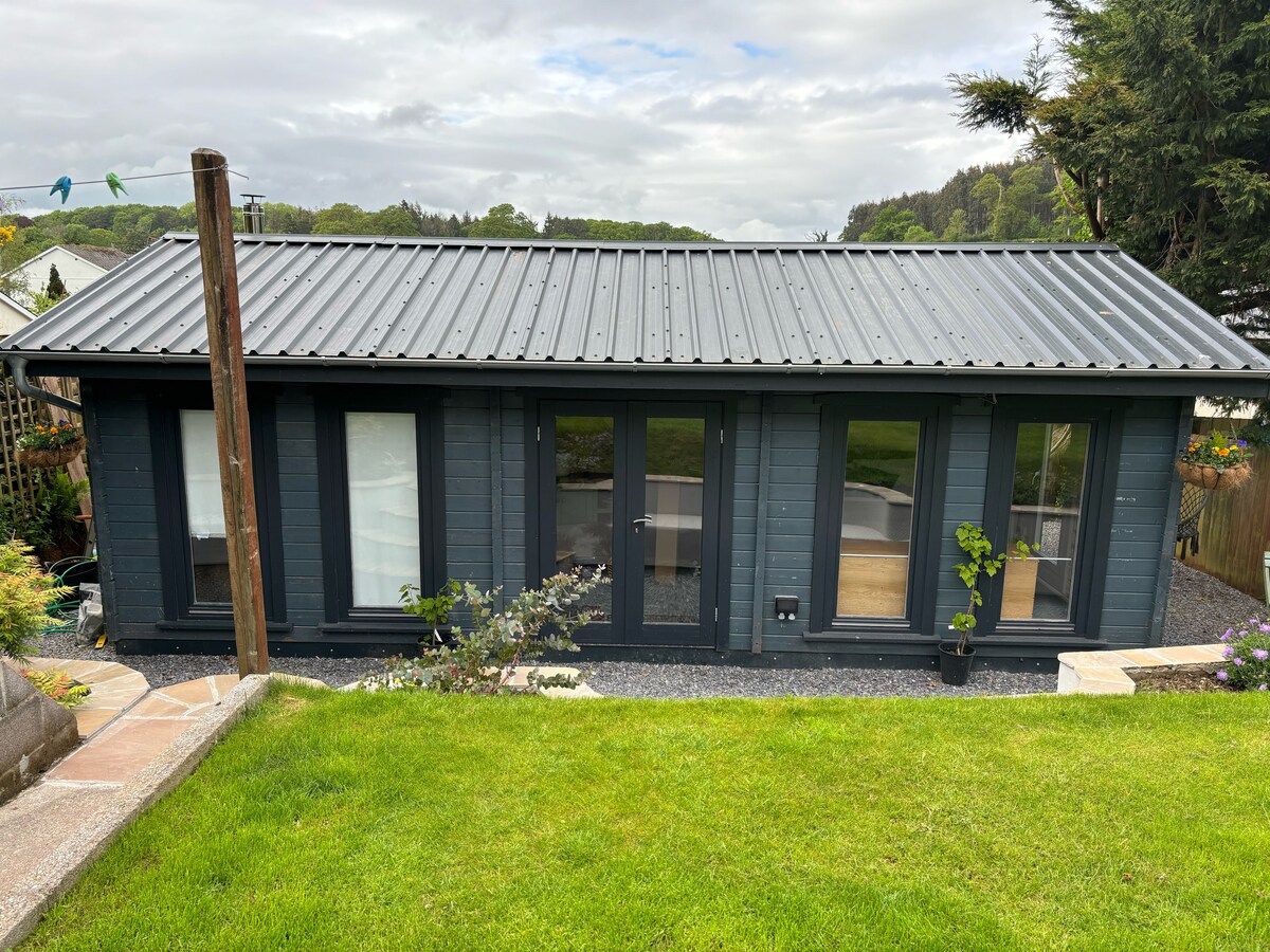 The cabin features a modern exterior with a dark-colored facade and a sleek metal roof. Large glass doors invite natural light and provide views of the surrounding green space. A landscaped area with grass and stones leads up to the entrance.