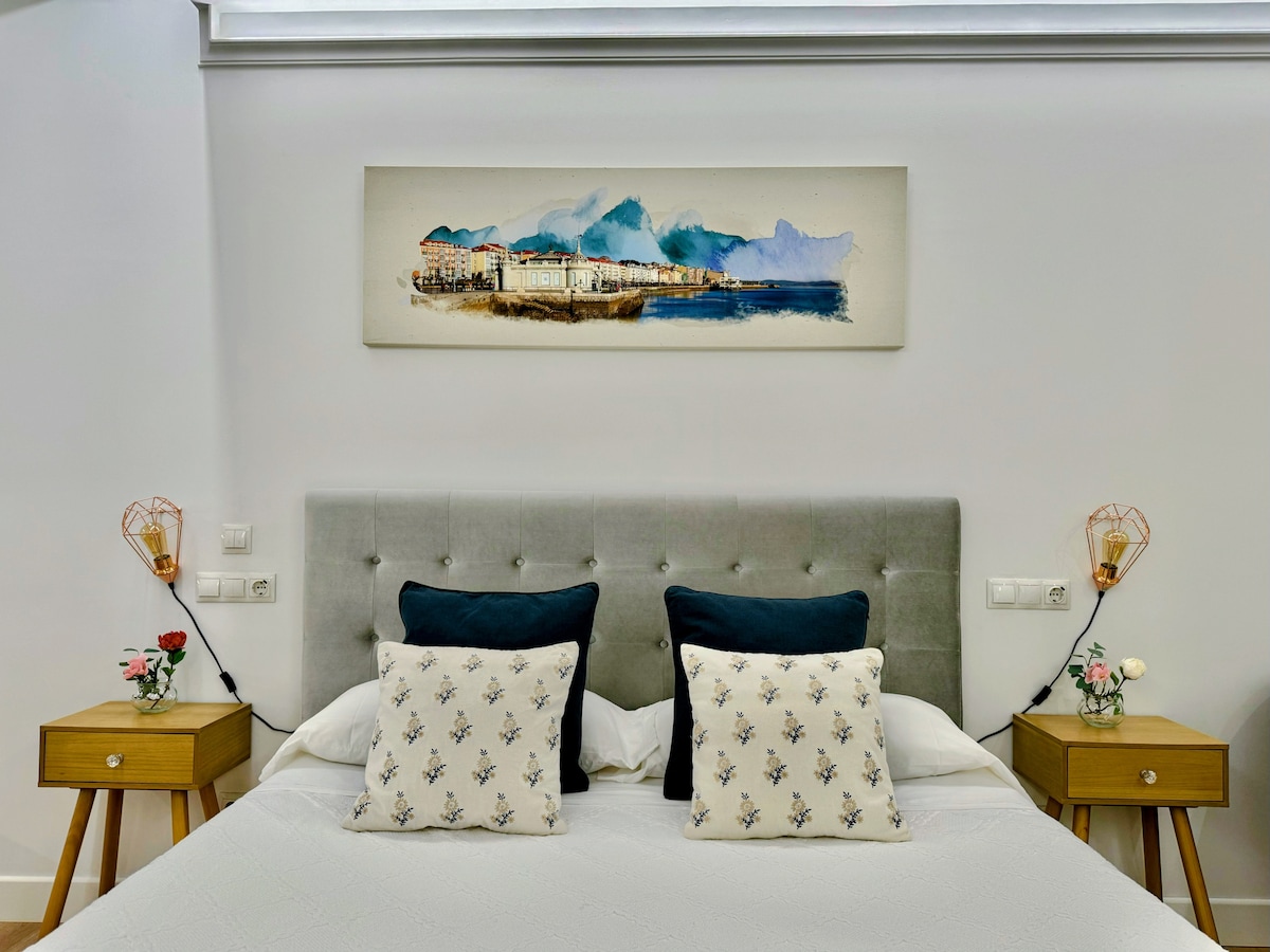 A spacious bed is centered in the room, flanked by two wooden nightstands with lamps. Decorative cushions add texture, and a coastal artwork hangs above the headboard, complementing the neutral color scheme of the space.
