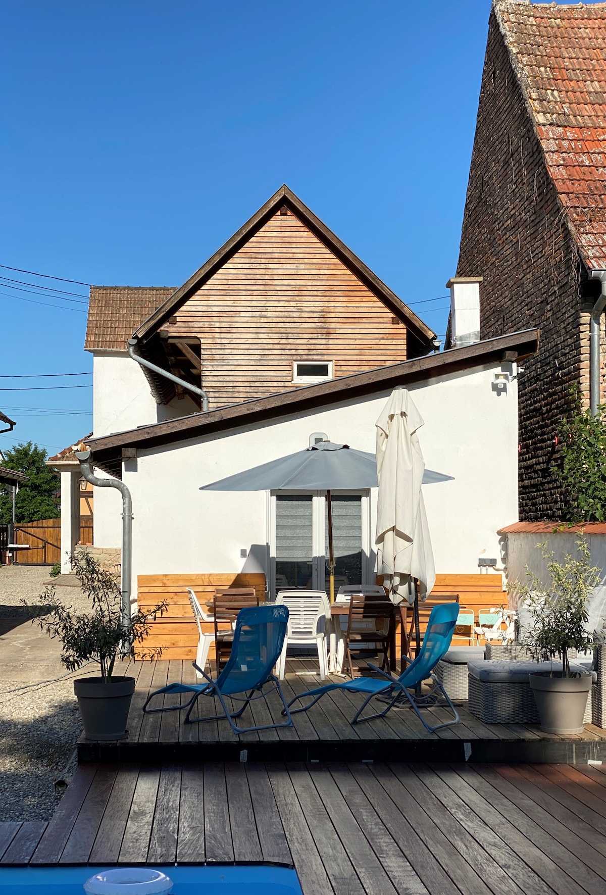A charming white house with a wooden roof is shown against a clear blue sky. Outdoor seating is arranged on the wooden deck, featuring a table, chairs, and an umbrella, creating a welcoming space for relaxation.