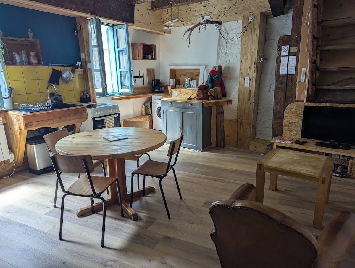 A bright kitchen and living area features wooden furnishings, including a round dining table surrounded by four chairs. A cozy armchair is positioned near a small television. Blue accents complement the yellow backsplash in the kitchen, which includes basic amenities and an open layout.