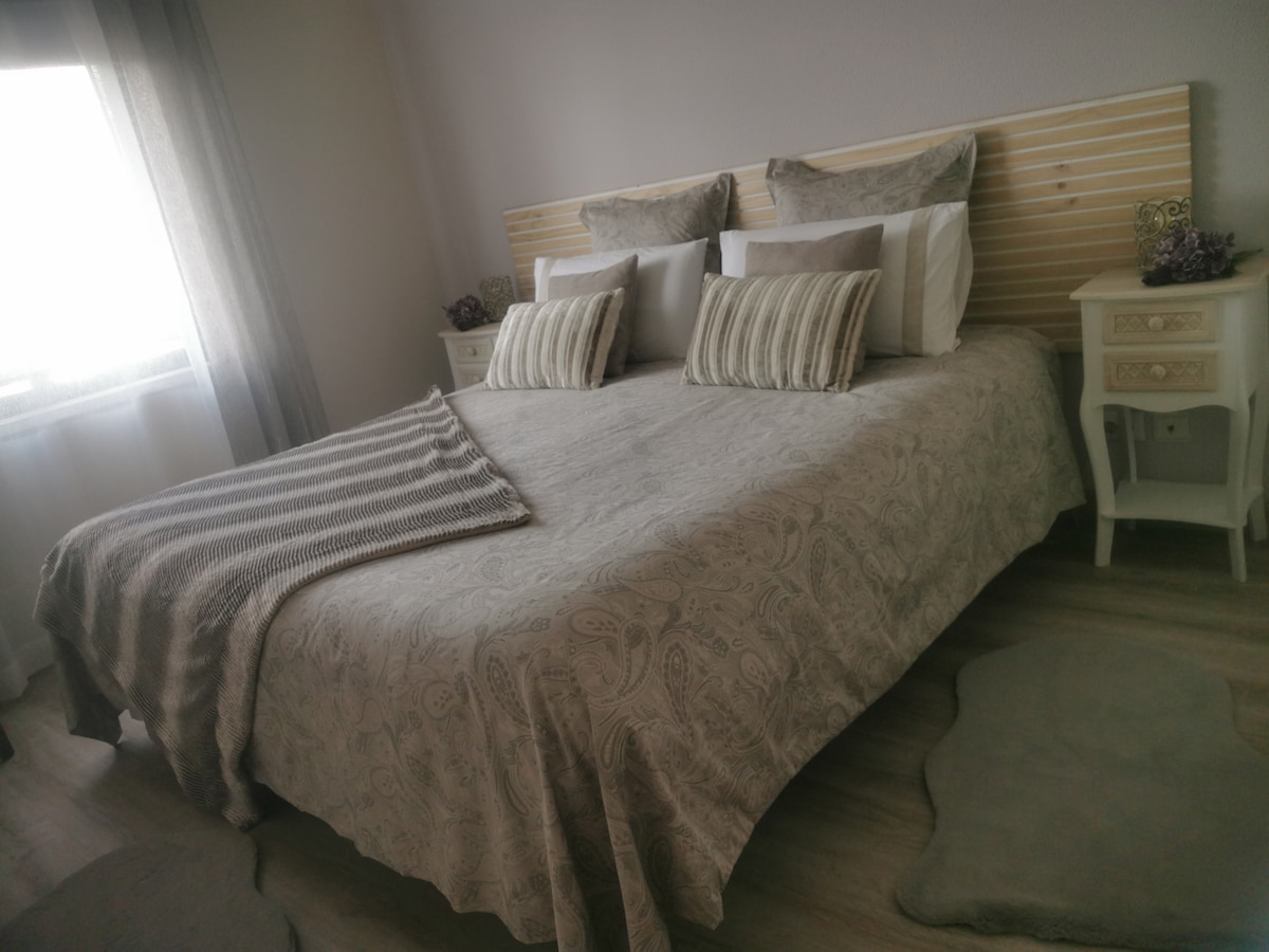 A spacious bed is dressed in soft bedding, flanked by nightstands with decorative accents. Light enters through a window with sheer curtains, creating a serene atmosphere. A textured throw is draped across the foot of the bed, and soft rugs are positioned on the floor.