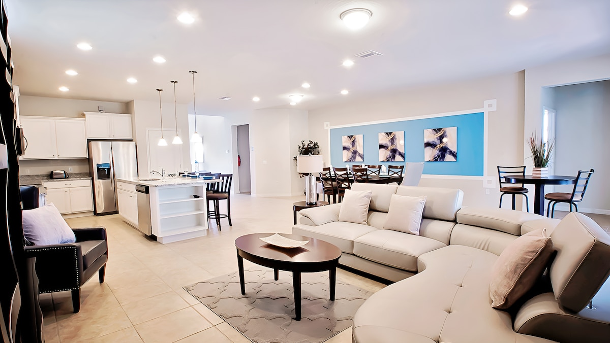 A spacious living area features a light-colored sectional sofa and a round coffee table on a soft rug. The dining area includes a table with seating for six, and the well-equipped kitchen is visible in the background, showcasing modern appliances and bright lighting.