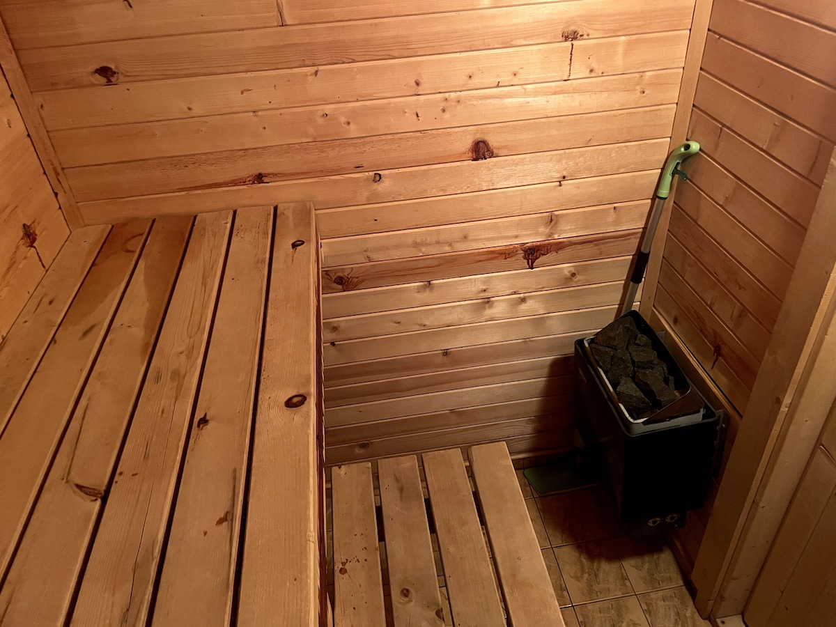 The interior of a wooden sauna is shown, featuring smooth wooden benches and paneling. A compact stove is positioned in the corner, with a small container of stones nearby. The warm wood tones create a calming environment suited for relaxation.