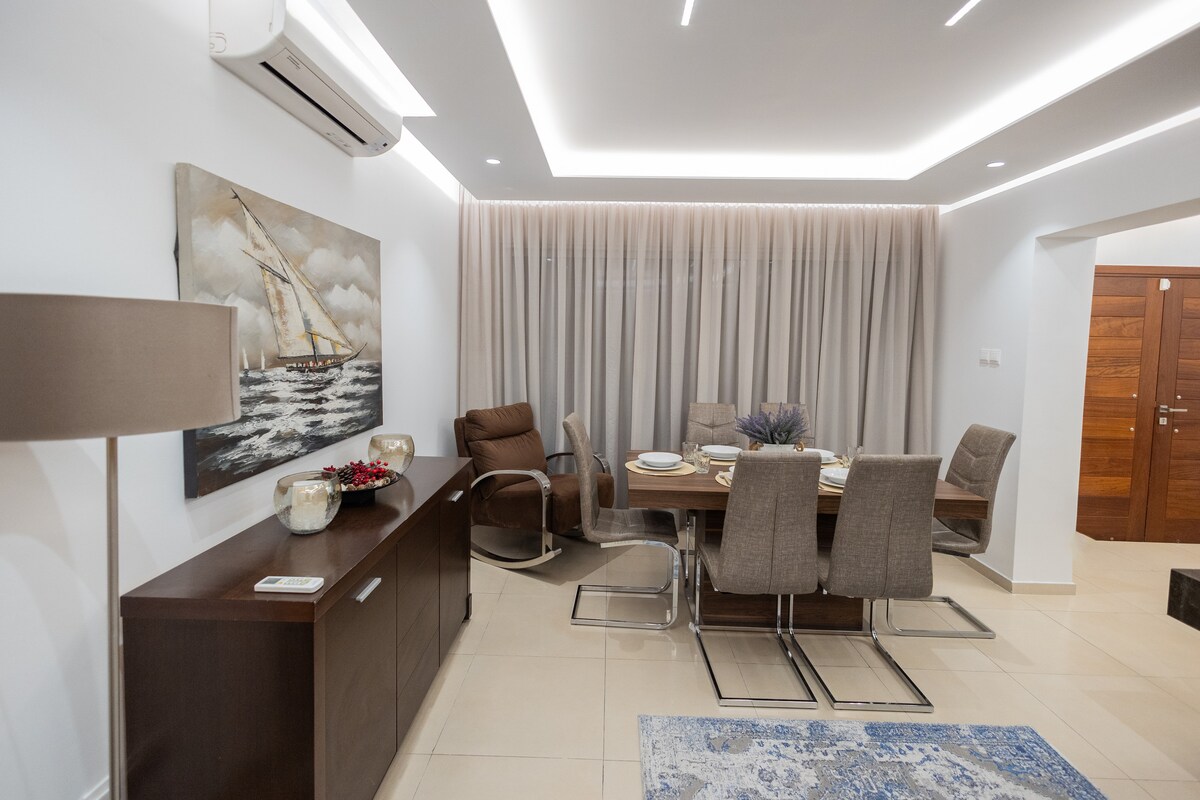 A dining area features a modern table surrounded by light-colored chairs. A painting of a sailboat adorns the wall, while a sideboard with decorative items is positioned nearby. Soft curtains drape across the windows, and an air conditioning unit is mounted above.