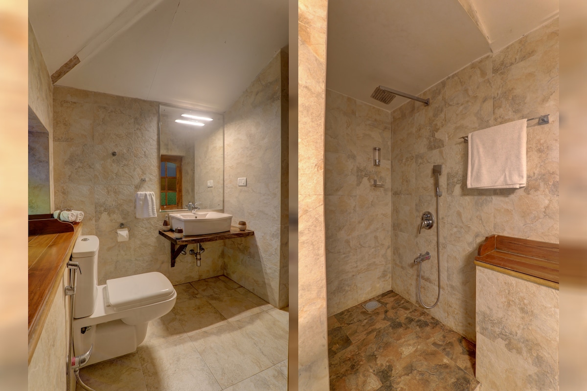 The bathroom features a modern layout with stone walls and a large window providing natural light. A washbasin is situated atop a wooden countertop, while a shower area is equipped with a rainfall showerhead. Towels are neatly arranged for guest convenience.