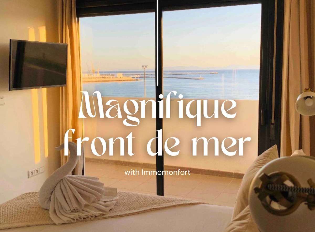 A spacious bedroom is presented with large glass doors opening to a sea view. Soft morning light filters through the curtains, illuminating the elegant decor. A television is mounted on the wall, and decorative towels are arranged attractively on the bed.