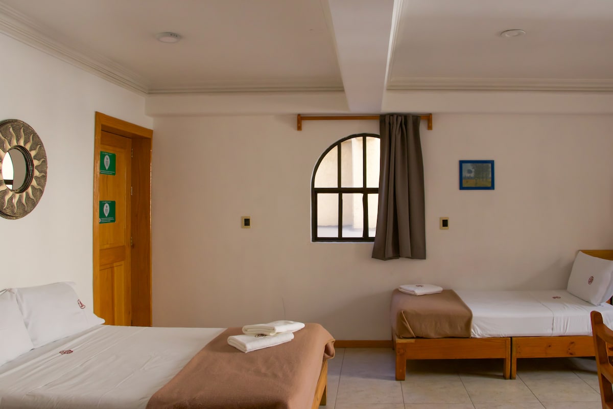 The room features two beds, one spacious and one single, positioned next to a window that allows natural light to enter. A round mirror hangs on the wall, complemented by a framed artwork. Neutral-toned decor contributes to a simple and comfortable ambiance.
