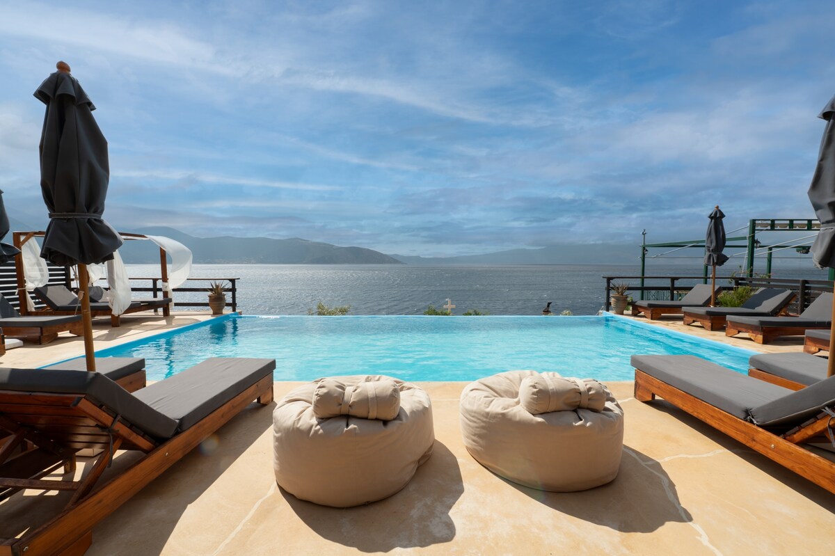 A tranquil pool area features comfortable lounge chairs arranged around a swimming pool, with plush bean bag seating situated close by. The serene view of the sea extends to the horizon, complemented by a backdrop of gentle hills under a partly cloudy sky.