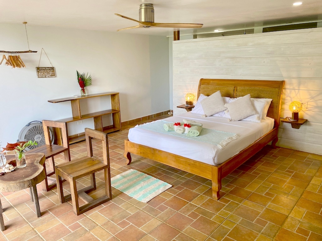 A bright room features a spacious bed with light linens and decorative pillows. Two wooden chairs and a small table are arranged nearby. A fan circulates air above, and tiled flooring enhances the natural feel of the space.