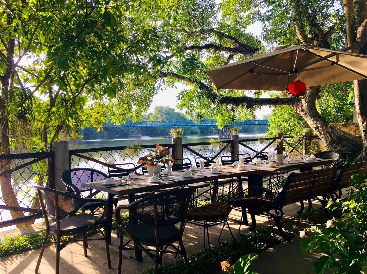 An outdoor dining area is surrounded by lush greenery, featuring a long table set with dishes and decor. An umbrella provides shade, and a river can be seen beyond the wooden railing, offering a serene morning setting.
